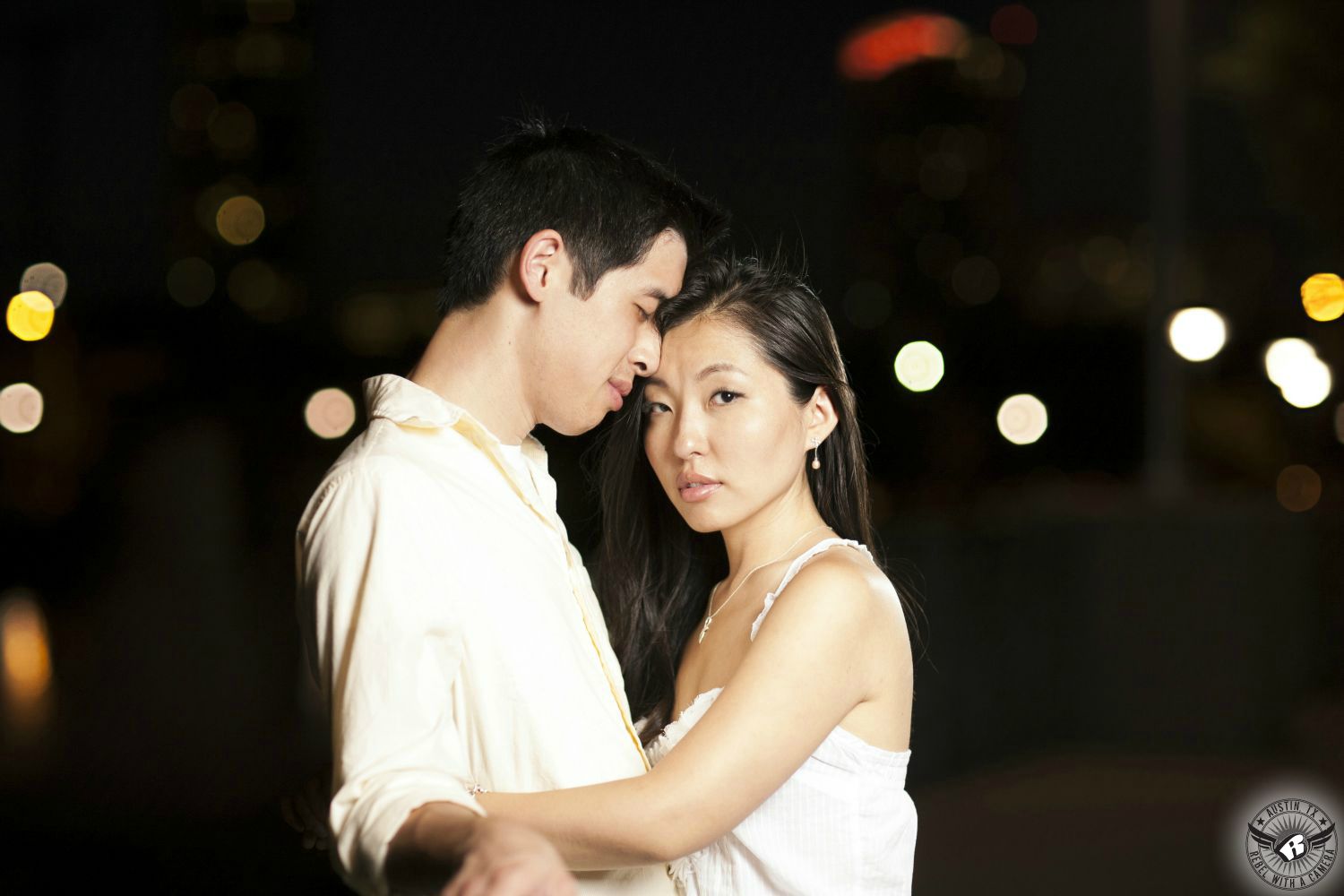 Sultry, Sexy, dark haired Chinese girl in a white spaghetti strap tank top leans against a Chinese guy with brown spiky hair wearing a yellow button up dress short with sleeves rolled up in the dark with bokeh spots of light in the background in this Austin inspired engagement portrait at The Palmer Events Center.